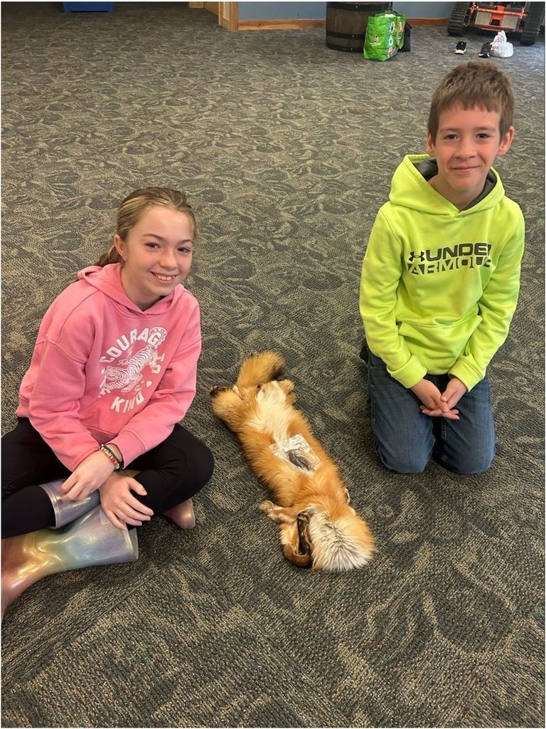 Two students sit on the floor beside a fox pelt, smiling at the camera while examining the specimen during a classroom activity.