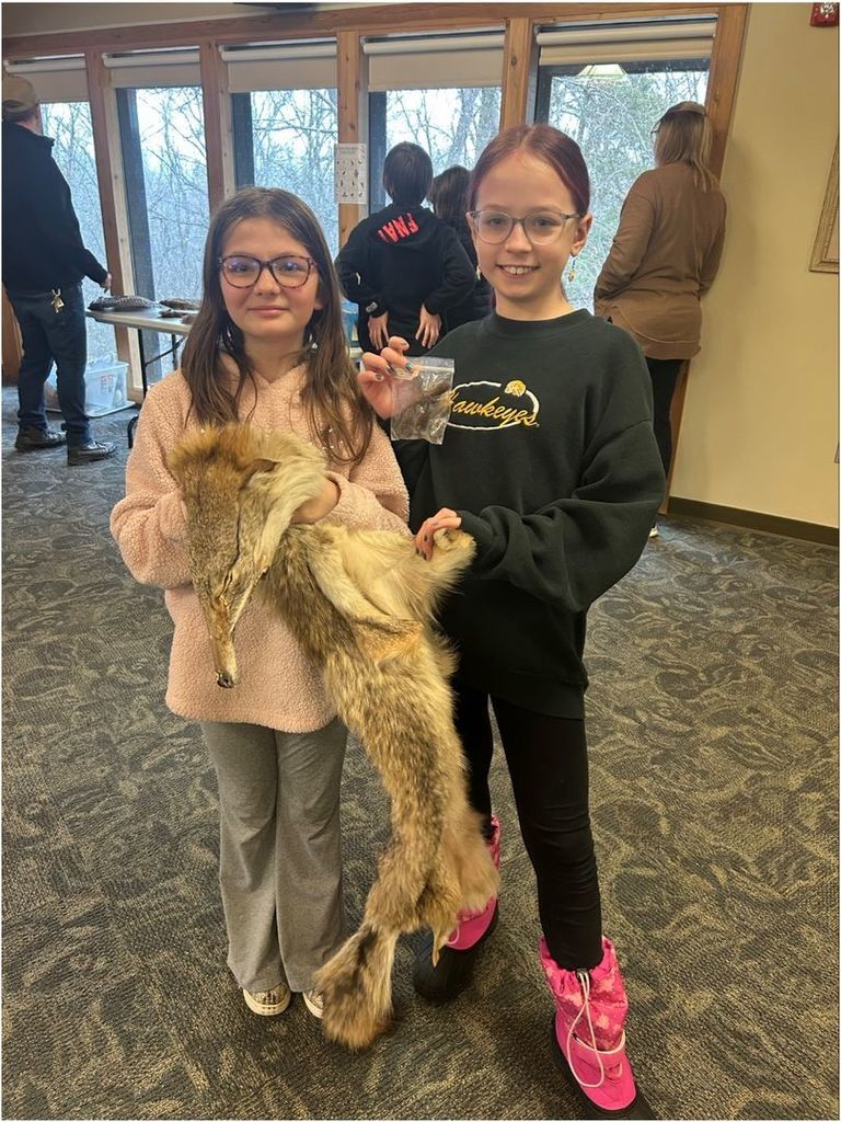 Two students hold a coyote pelt and a small sample bag, smiling while standing in a classroom with large windows and wooded scenery outside.