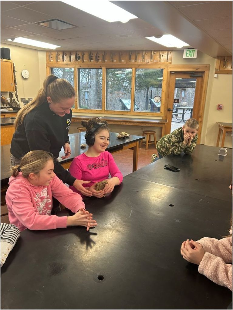 Students gather around a table as an instructor guides one student holding a turtle shell; others watch with curiosity in a nature classroom.