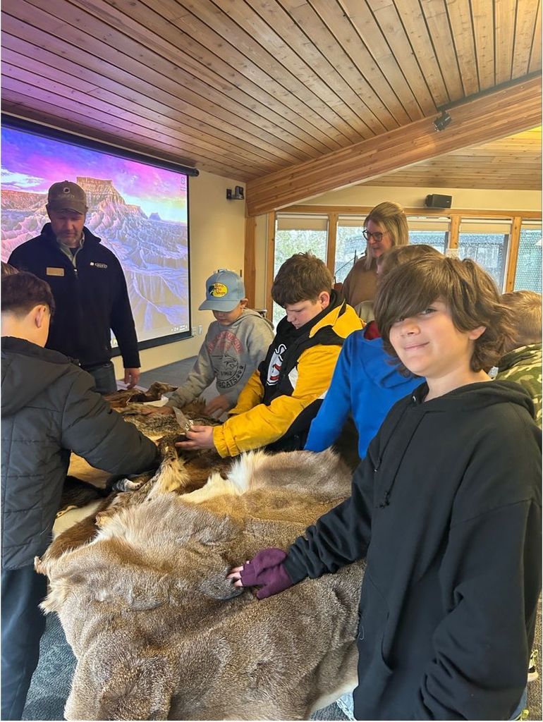 Students gather around a table covered with animal pelts, touching and examining them while an instructor supervises in a learning space.