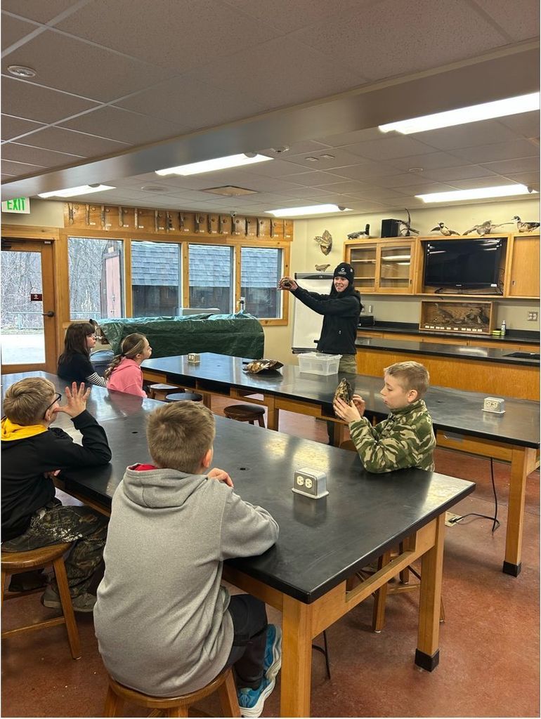 Instructor holds up a turtle shell while students sit at lab tables, raising hands and observing during a hands-on science lesson.