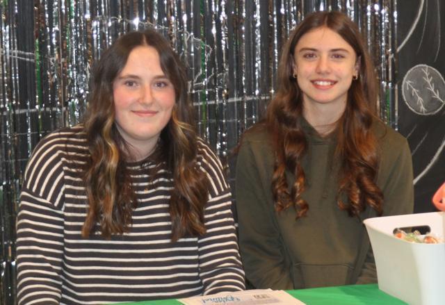 Two girls sit side by side, smiling at a table with papers, in front of a sparkly backdrop at a school event.