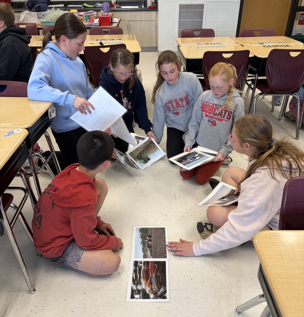Six students sit in a circle, holding and comparing printed photos spread on the classroom floor.