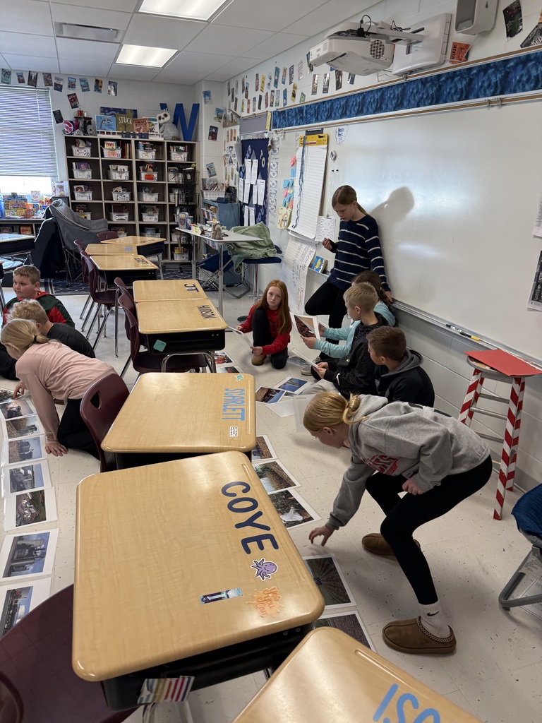 Students kneel on a classroom floor arranging printed photos in a line while a teacher observes nearby.
