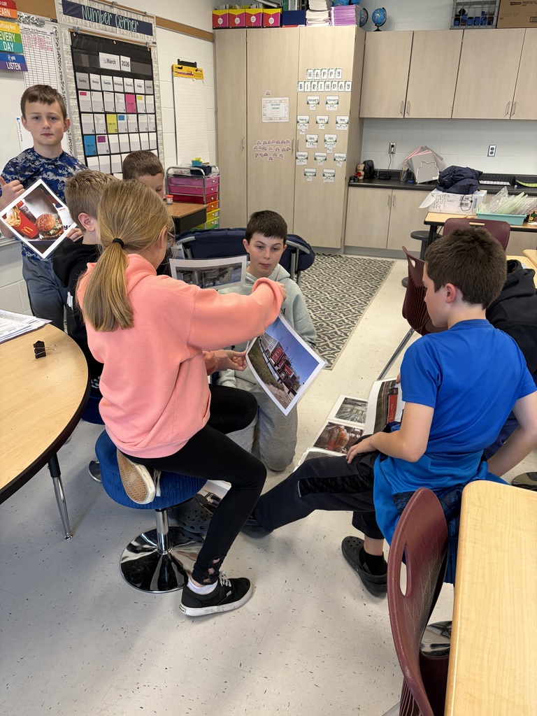 Students cluster around desks while a teacher guides them in reviewing a line of printed images on the floor.