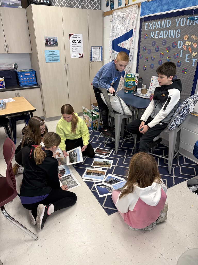 Students sit and kneel around a rug, sorting and discussing printed photos during a small-group classroom task.
