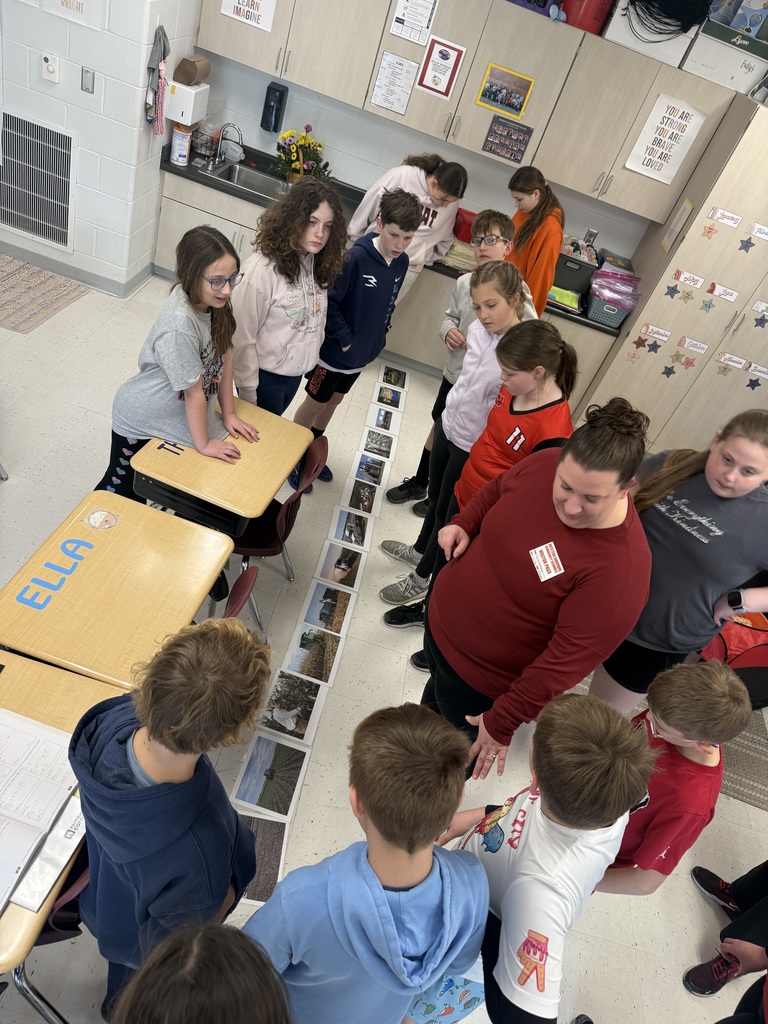 Students cluster around desks while a teacher guides them in reviewing a line of printed images on the floor.