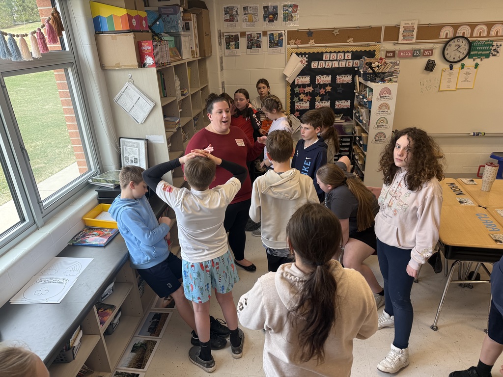 A teacher speaks to a group of students gathered near a window, with printed images laid out along the floor.