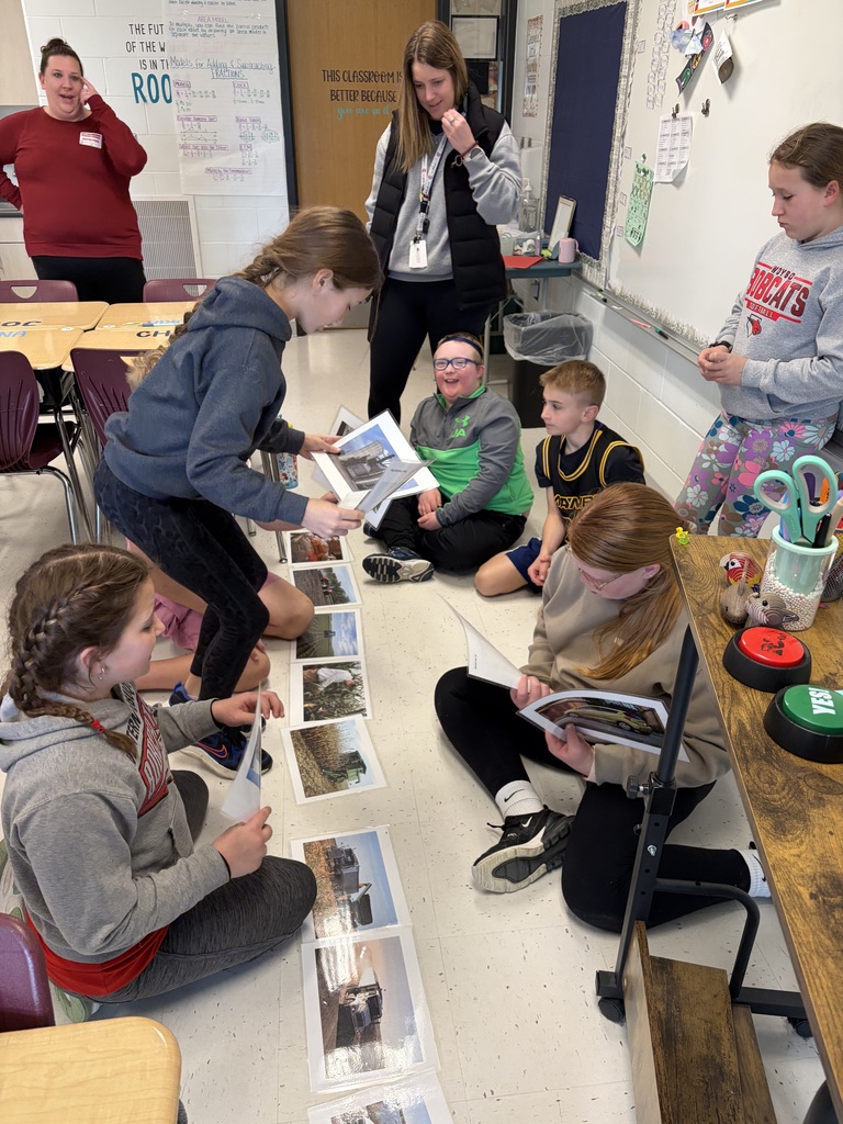 A teacher stands with students gathered around as one student shares printed photos during a classroom activity.