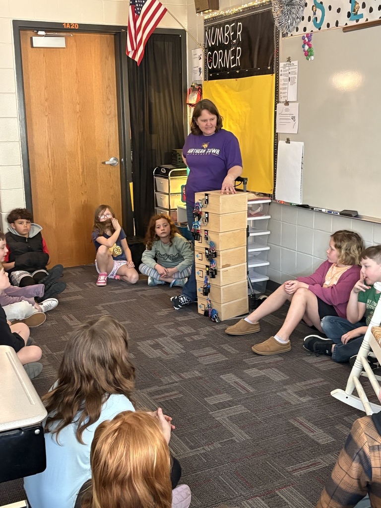 A teacher sits with students in a circle, explaining a breakout box activity while students listen attentively.