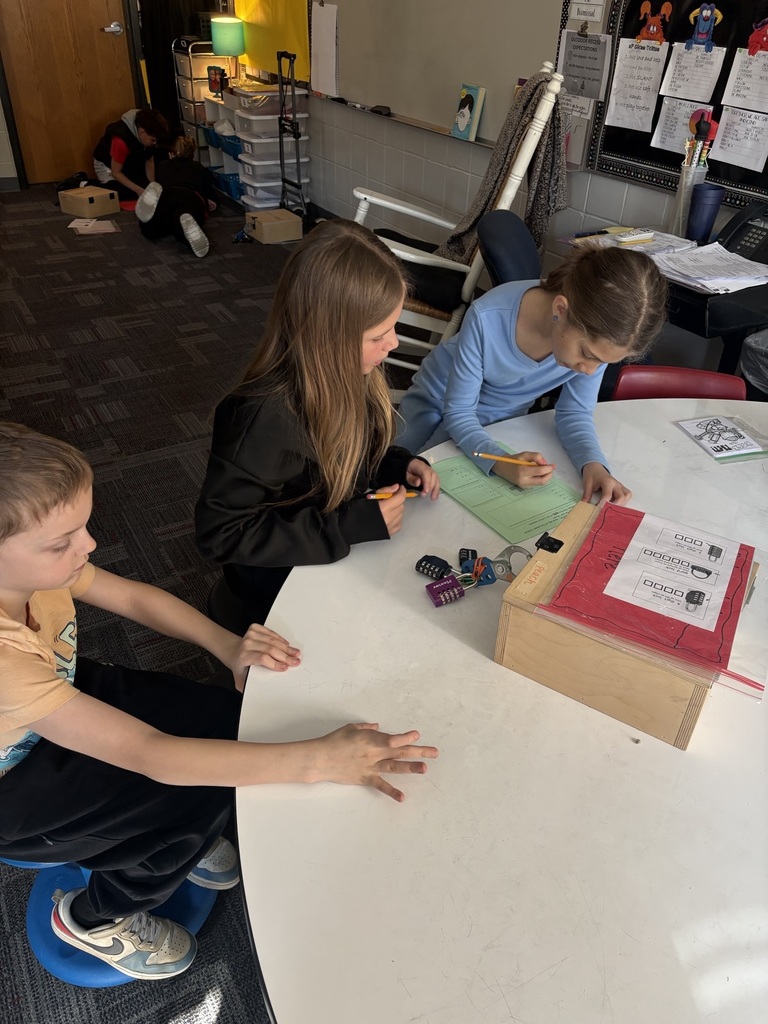 Three students sit at a table, writing on a worksheet while working with locks and a breakout box activity.