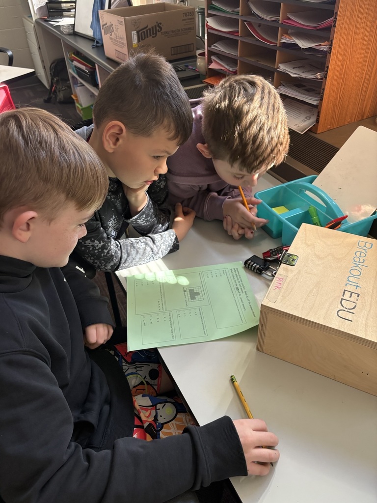Three students lean over a worksheet, studying clues and tools beside a breakout box during a group challenge.