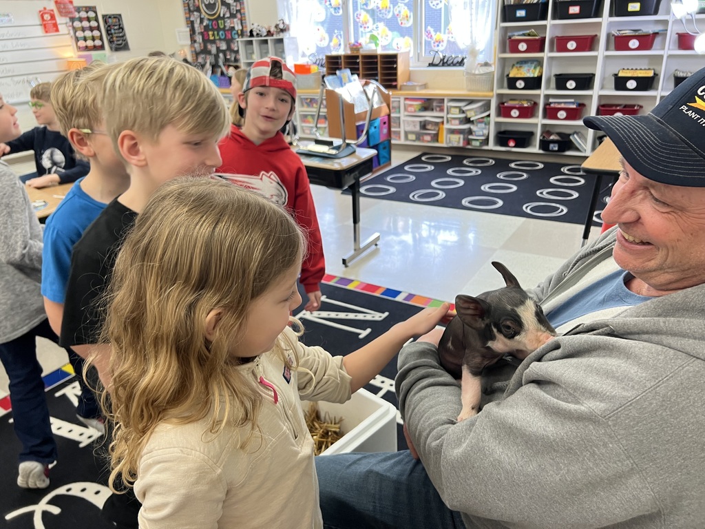 Young student smiles while gently petting a small pig held by an adult, with classmates nearby.