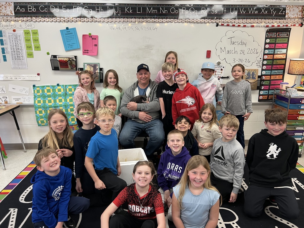 Class group photo with students and an adult holding a small pig, all smiling in a decorated classroom.