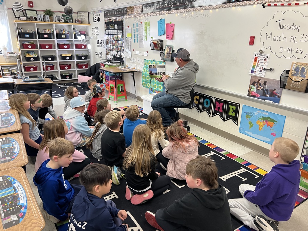 Adult reads aloud to seated elementary students on a classroom rug during a group lesson.