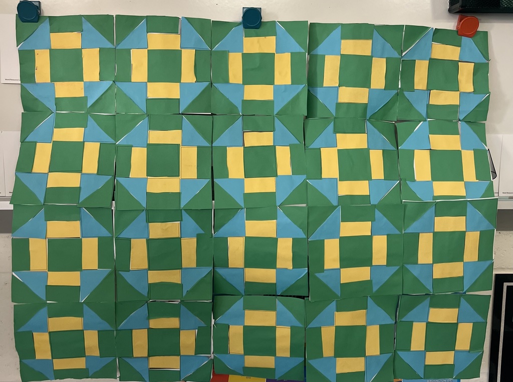 Display of colorful paper quilt squares arranged in a repeating geometric pattern on a classroom wall.