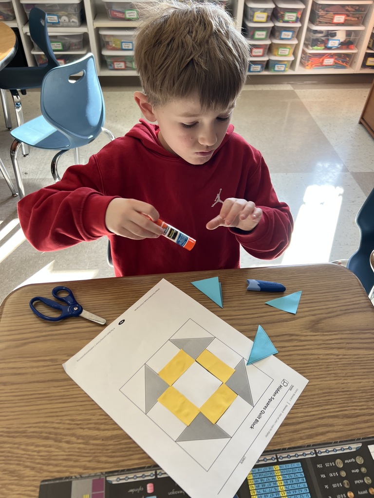Student applies glue to paper pieces while building a patterned quilt design on a worksheet at their desk.