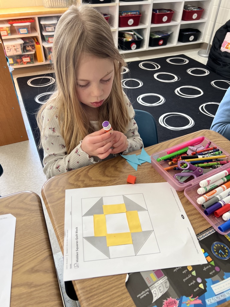 Student glues paper shapes onto a template, assembling a geometric quilt pattern with scissors and supplies nearby.