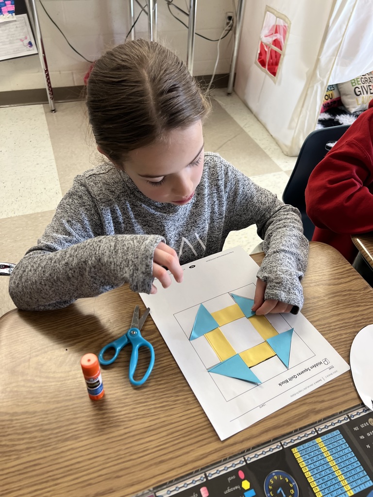 Student glues paper shapes onto a template, assembling a geometric quilt pattern with scissors and supplies nearby.