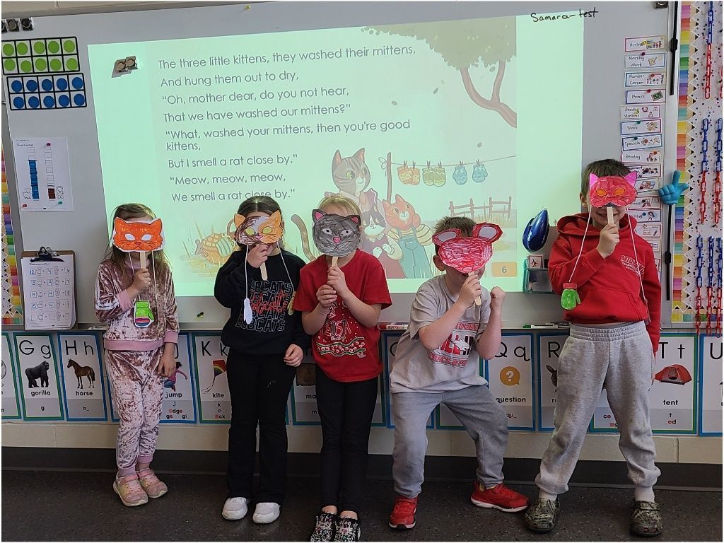 Five students pose playfully with colorful animal masks and props while acting out a classroom reading activity in front of the board.