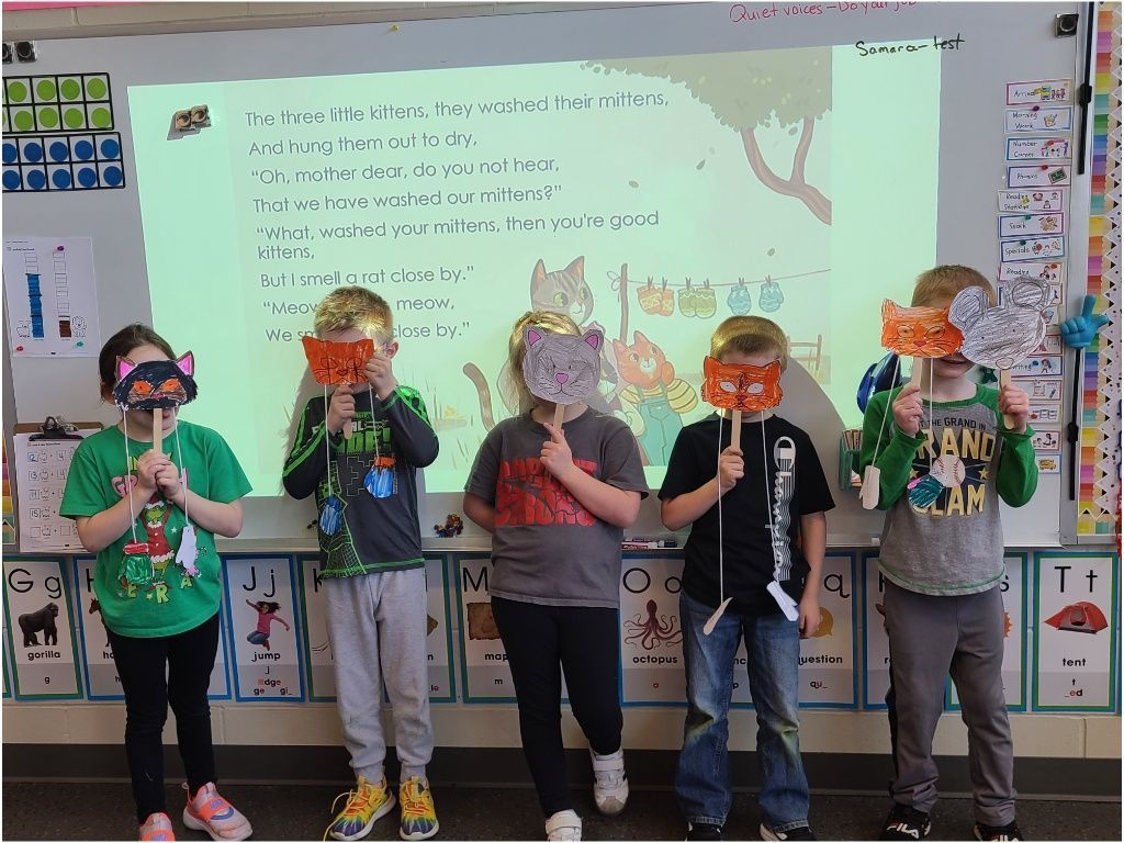 Five students stand in a row holding animal masks over their faces while participating in a reading activity about “Three Little Kittens.”