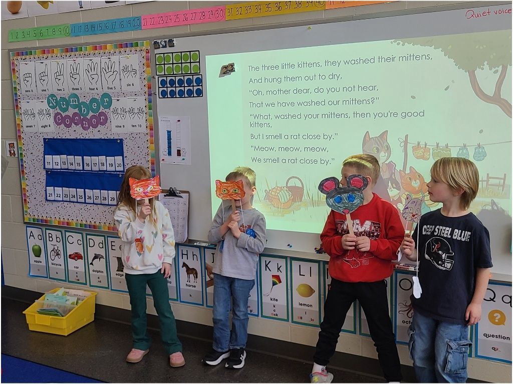 Four students hold handmade cat masks on sticks, standing in front of a classroom screen displaying the “Three Little Kittens” rhyme.