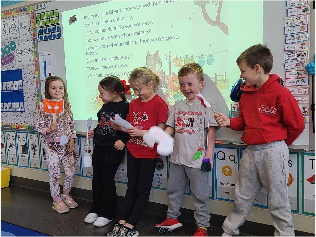 Five students stand together smiling and holding paper props and masks as they act out parts of the “Three Little Kittens” poem.