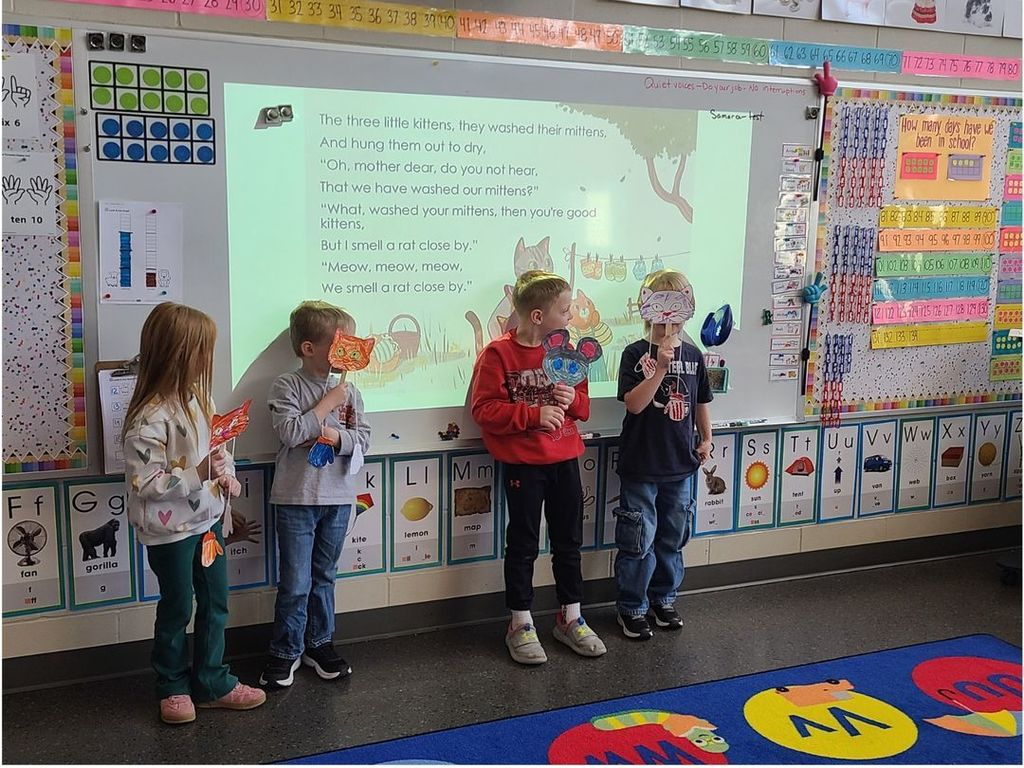 Four young students stand at the front of a classroom holding colorful animal masks while following along with a projected “Three Little Kittens” poem.