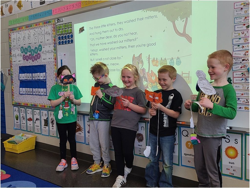 Five students smile and hold up decorated animal masks during a classroom activity in front of a projected story.