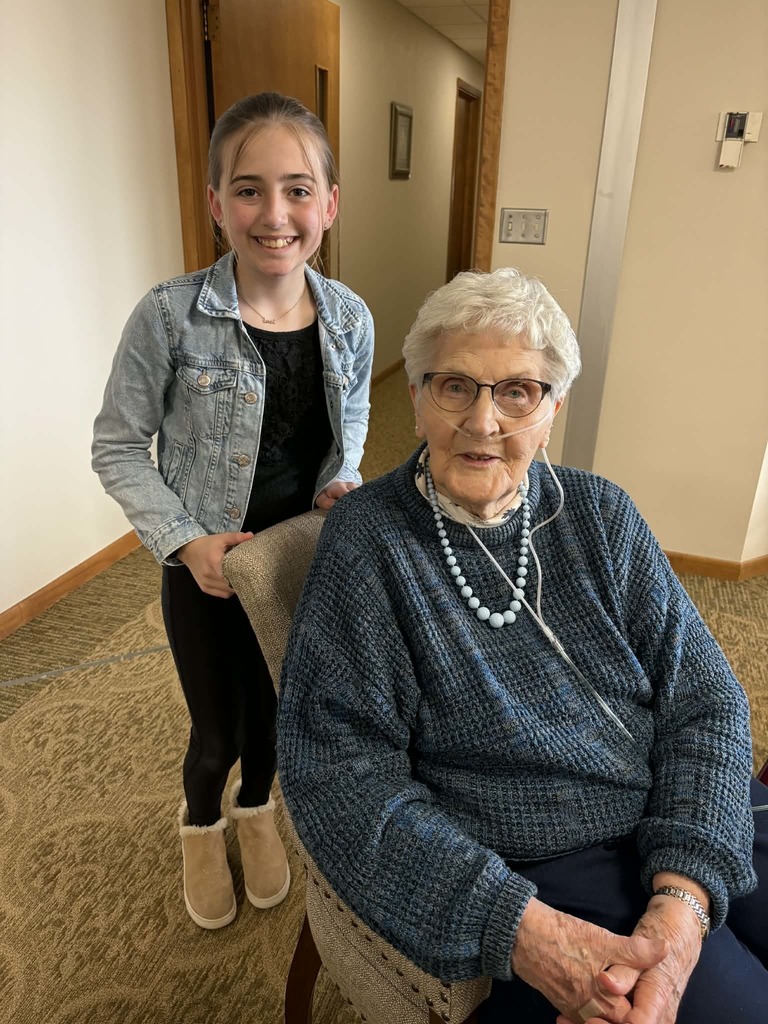Girl stands behind an elderly woman seated in a chair, both smiling gently in a hallway setting.
