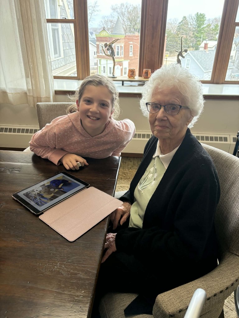Girl and elderly woman sit by a window with a tablet on the table, sharing a moment together in a bright room.