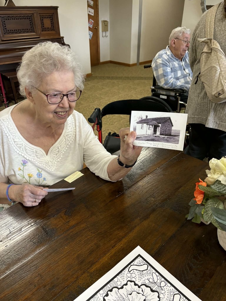 Elderly woman holds up an old black-and-white photo at a table, sharing a memory in a social gathering space.