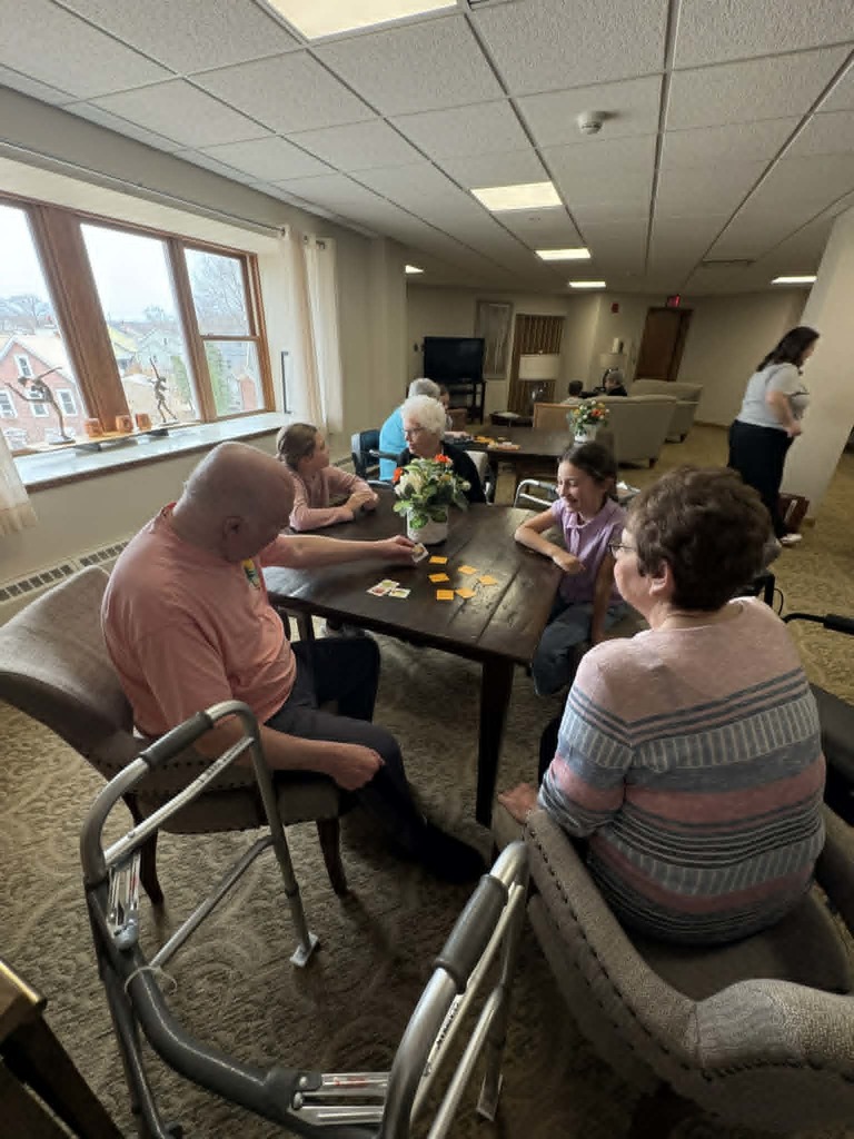 Group of children and older adults gather around tables playing games in a bright, welcoming community room.