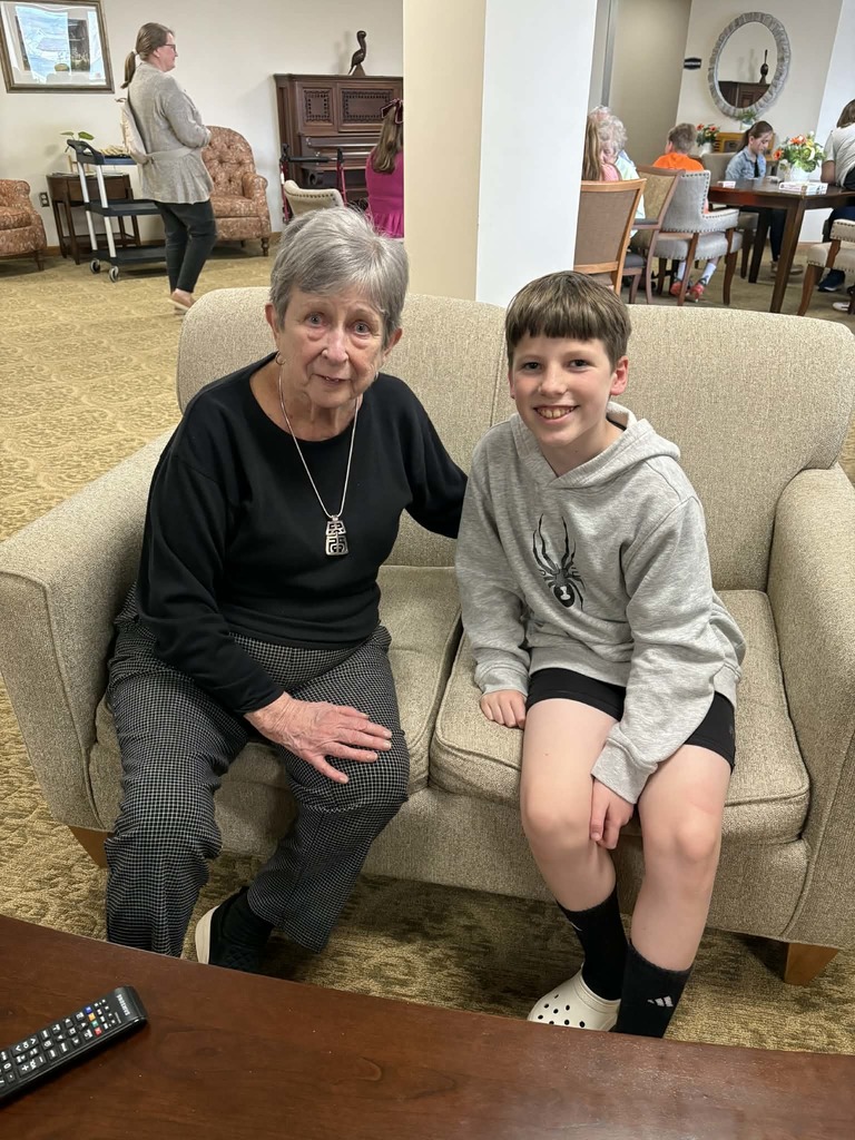 Elderly woman and boy sit together on a couch, smiling warmly in a comfortable common area.