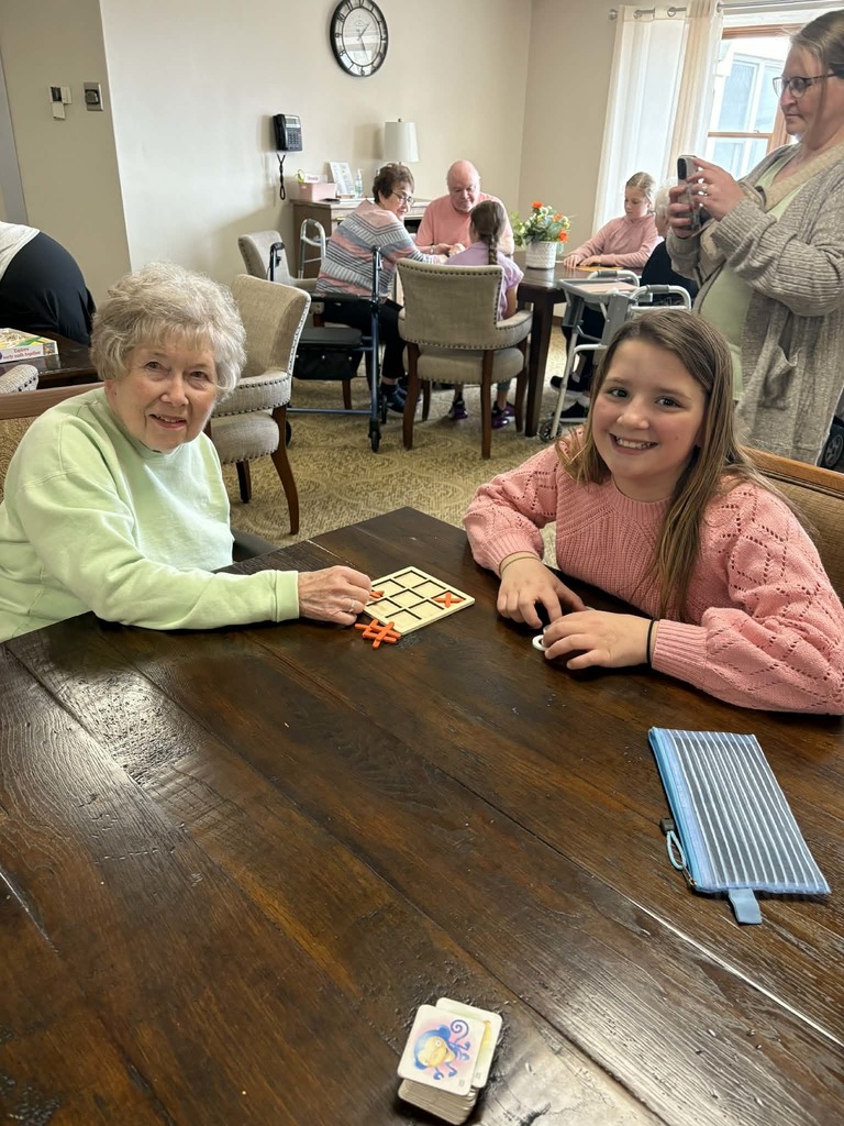 Elderly woman and girl play a tabletop game while others sit and talk in the background of a shared living space.