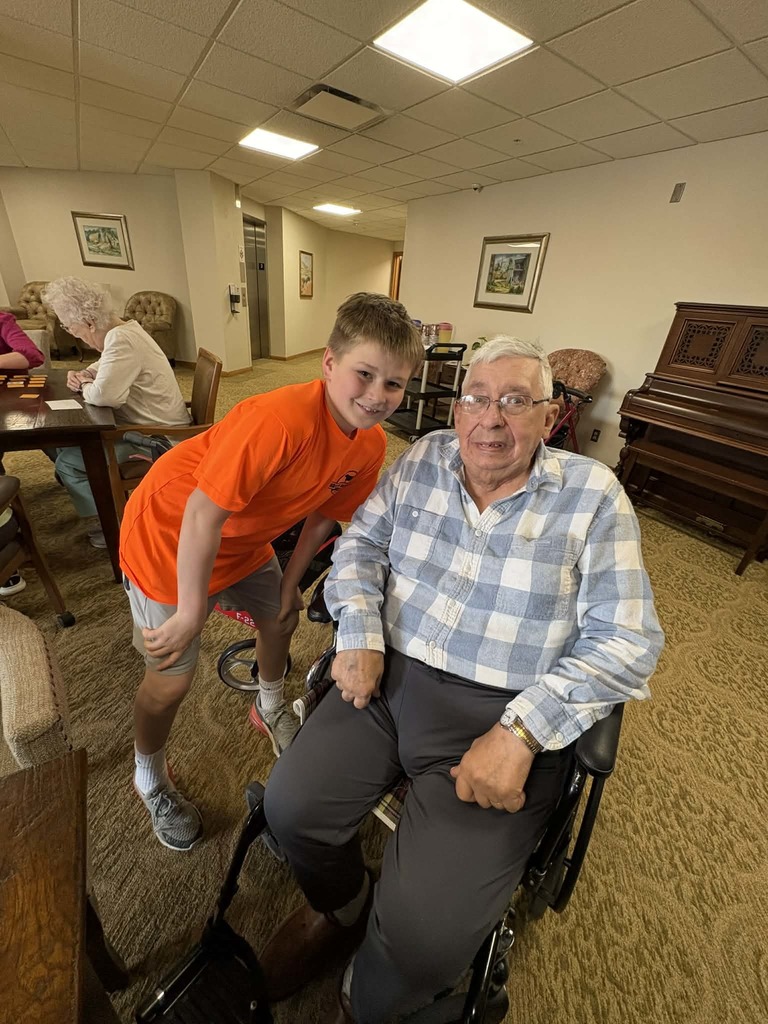 Boy leans beside an older man in a wheelchair, both smiling in a communal room with a piano in the background.