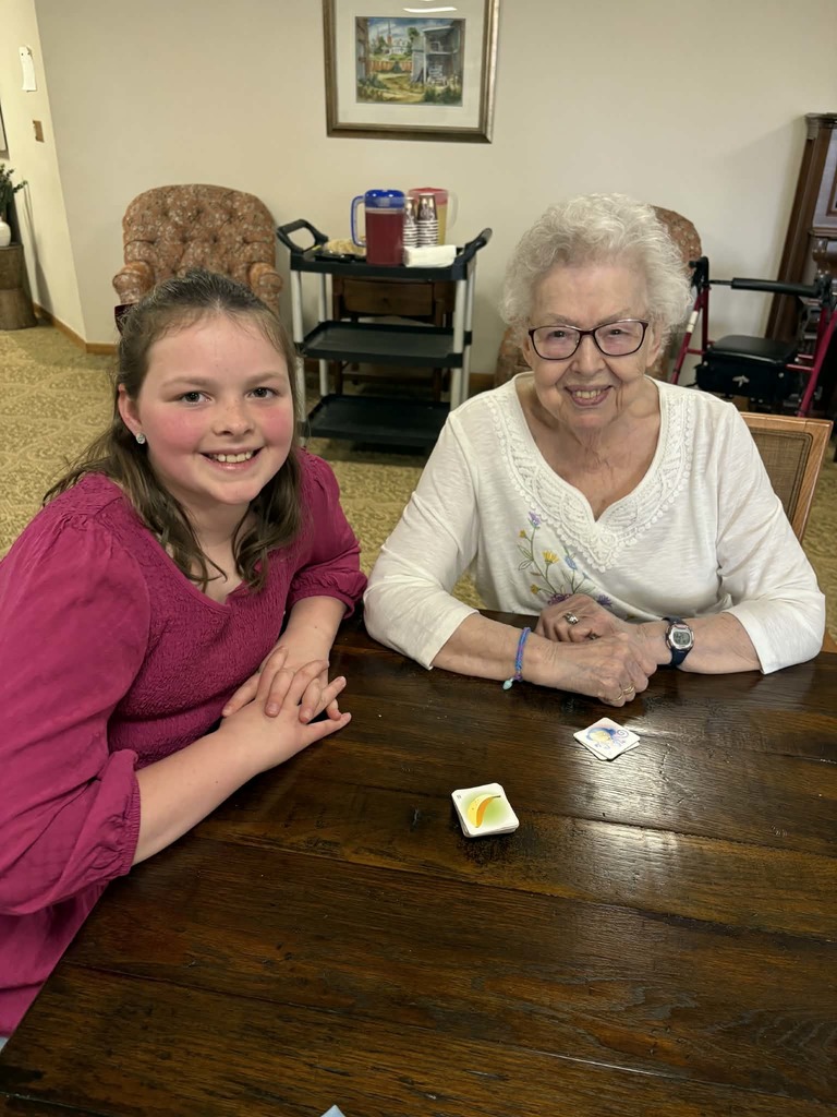 Young girl and elderly woman smile at a table with a few game cards between them in a cozy common room.