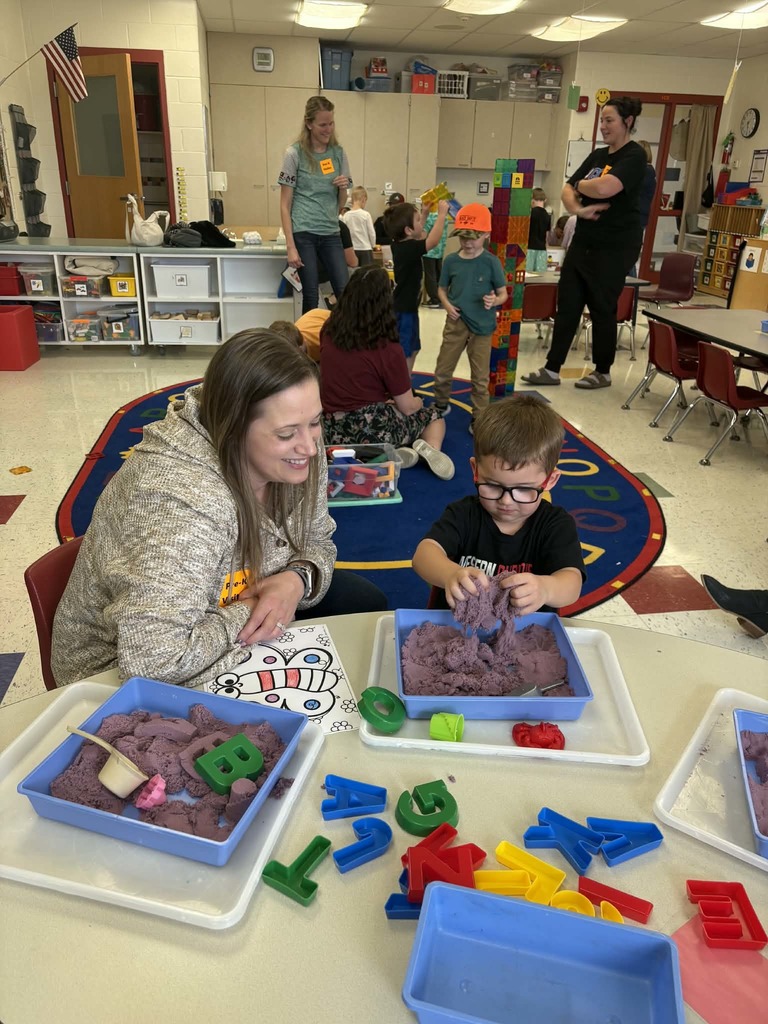 Child plays with purple kinetic sand and letter molds at a table while an adult smiles beside him in a busy classroom.