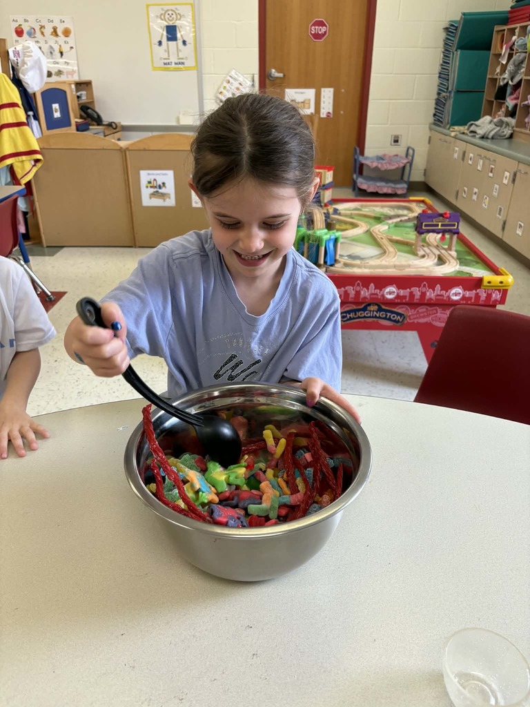 Young girl smiles while scooping colorful gummy candy from a metal bowl at a classroom table.