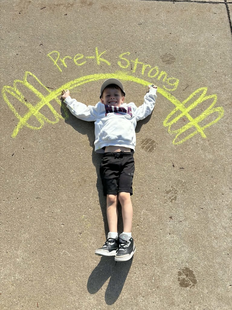 Child lies on pavement under chalk barbell drawing labeled “Pre-K Strong,” smiling with arms raised.