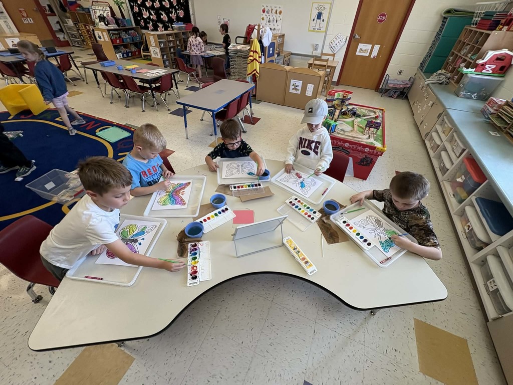 Children sit around a table painting butterfly pictures with watercolor sets in a classroom.