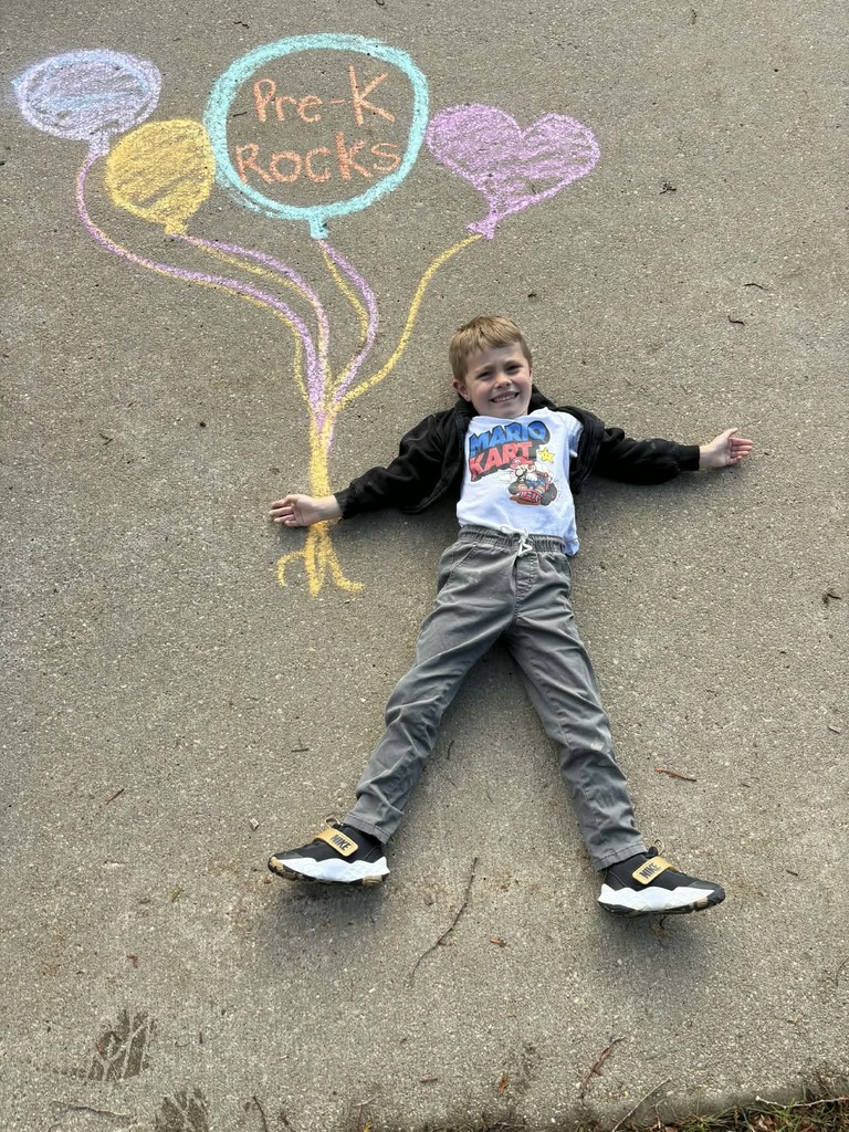 Child lies on pavement smiling beside chalk drawings of balloons with the words “Pre-K Rocks.”