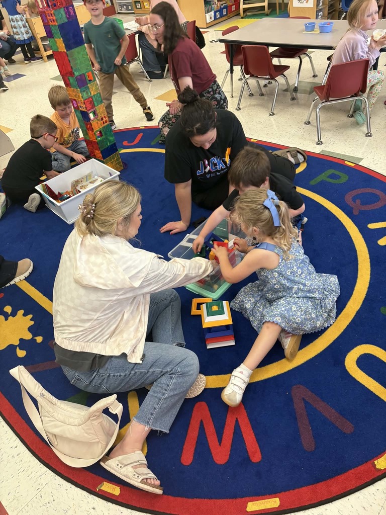 Children and adults sit on a classroom rug building with blocks, including a tall colorful tower nearby.