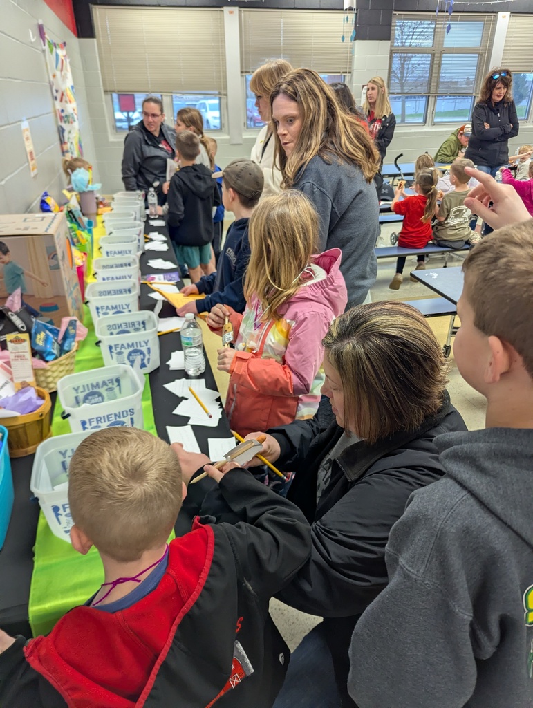 Families and children gather around a table with labeled bins, filling out slips during a school event.