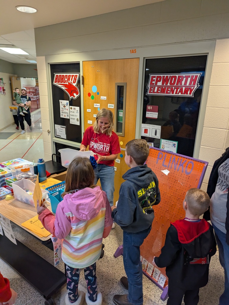 Students line up at a school table activity with a “Plinko” board while a staff member hands out materials in a hallway.