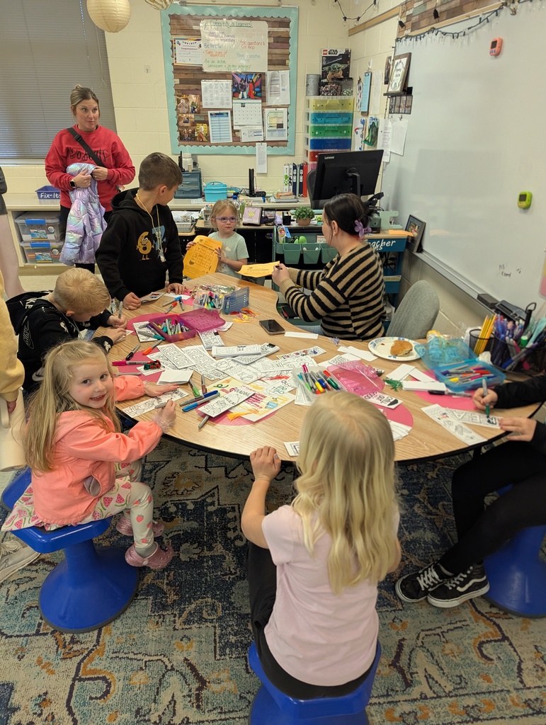Children and an adult sit around a classroom table coloring worksheets and crafting, with markers and supplies spread out.