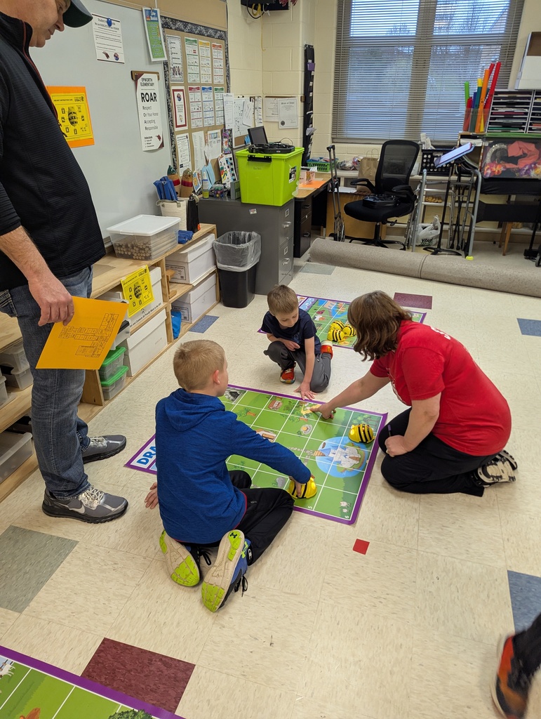 Teacher kneels with three children using a floor grid activity mat while another adult observes nearby in a classroom.