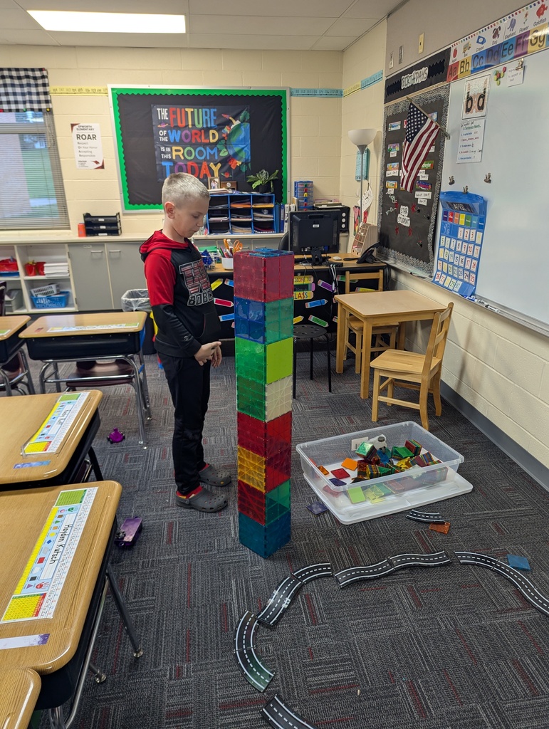 Child stands beside a tall tower built from colorful magnetic tiles in a classroom with bins of blocks nearby.