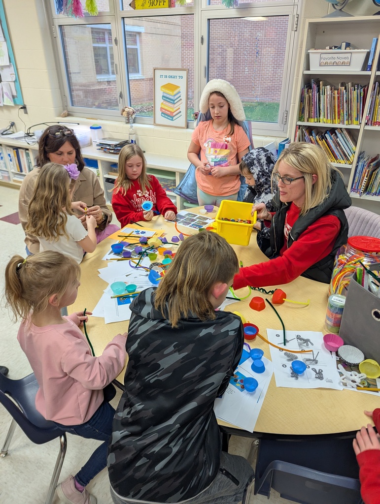Adults and children work together on a craft project at a classroom table using colorful materials like pipe cleaners and cups.