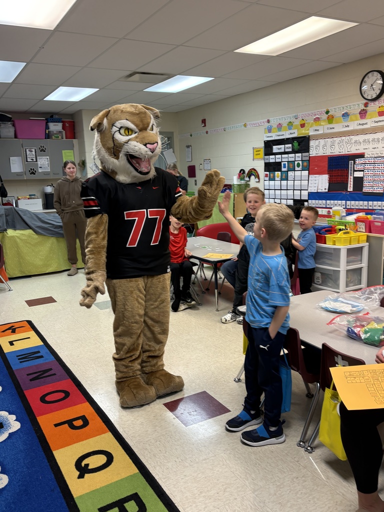 School mascot in a jersey gives a high-five to a smiling child while other students watch in a classroom.